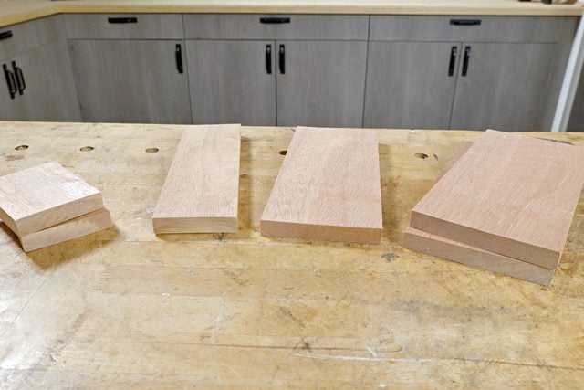 Four wooden planks of varying sizes are arranged side by side on a wooden workbench. In the background, gray cabinets with black handles are visible.