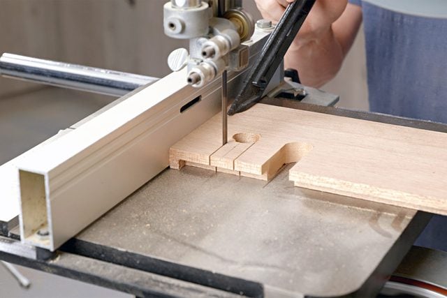 A person is cutting a piece of wood with a band saw, focusing on precise details. The wood is positioned against a guide fence for accuracy. The scene is likely set in a workshop or carpentry space.