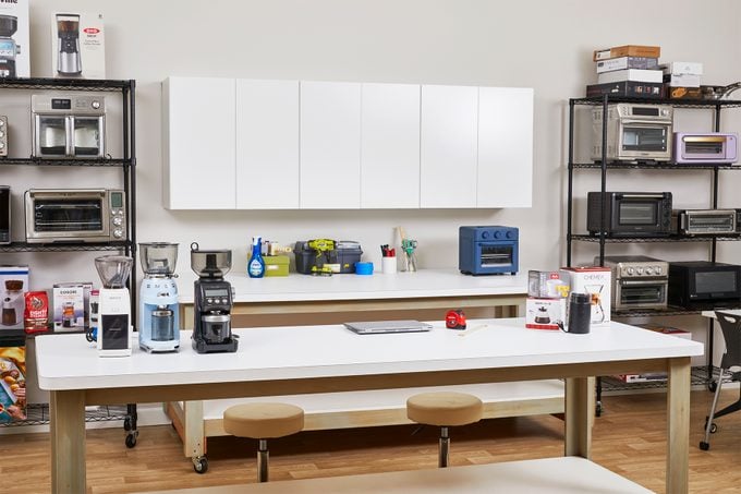 A well-organized kitchen showroom featuring various countertop appliances, including blenders, coffee makers, and toasters, displayed on white counters and metal shelves. The room has two large tables with stools in the center and wooden flooring.